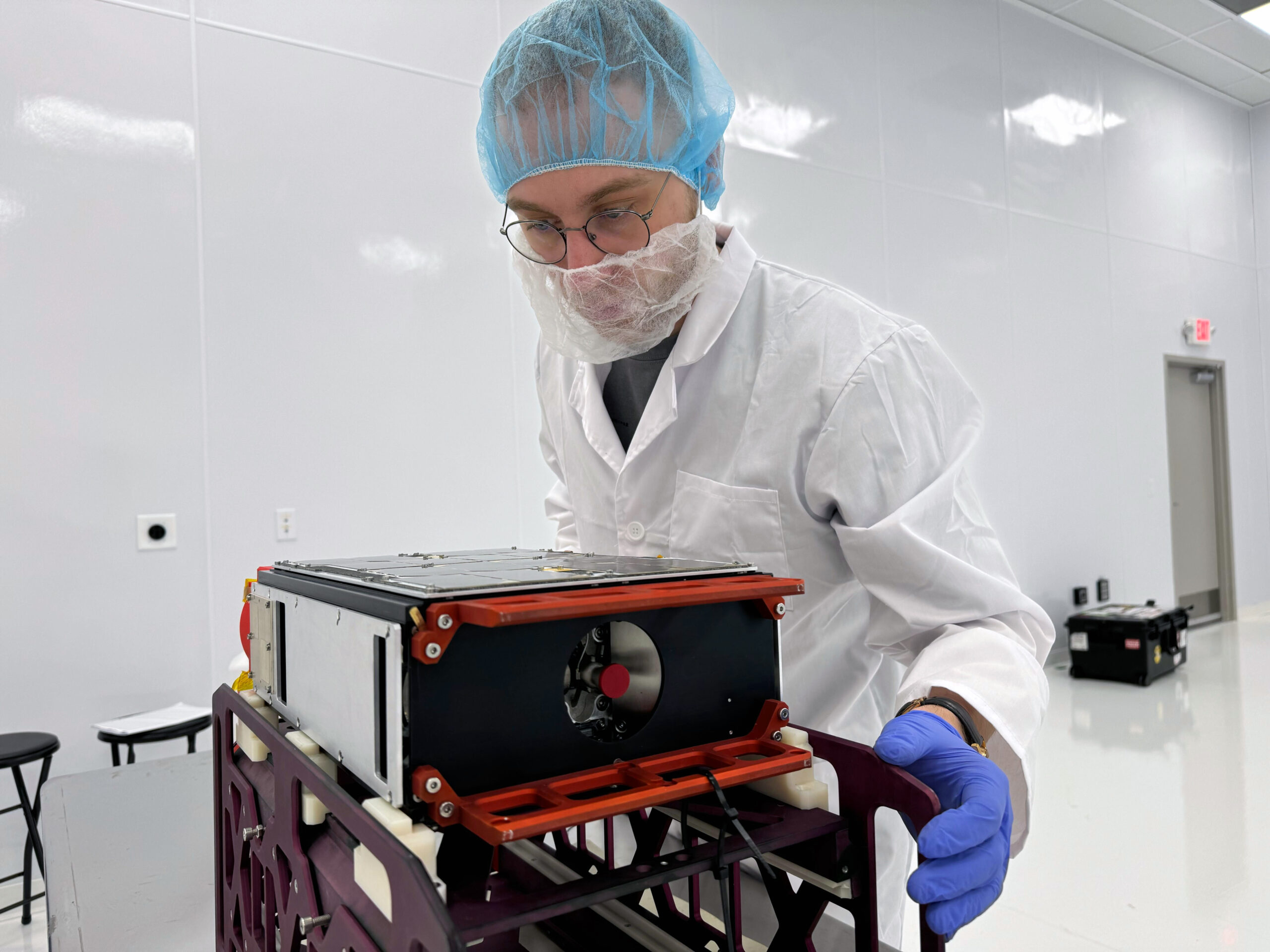 Lifteor enigneer in a cleanroom at Impulso Space facility in Melbourne, Florida, USA, inspects the RED5 satellite before the refueling process. The satellite is mounted on a support structure, and the engineer is wearing a lab coat, gloves, and a hairnet for contamination control. Credit: Liftero.